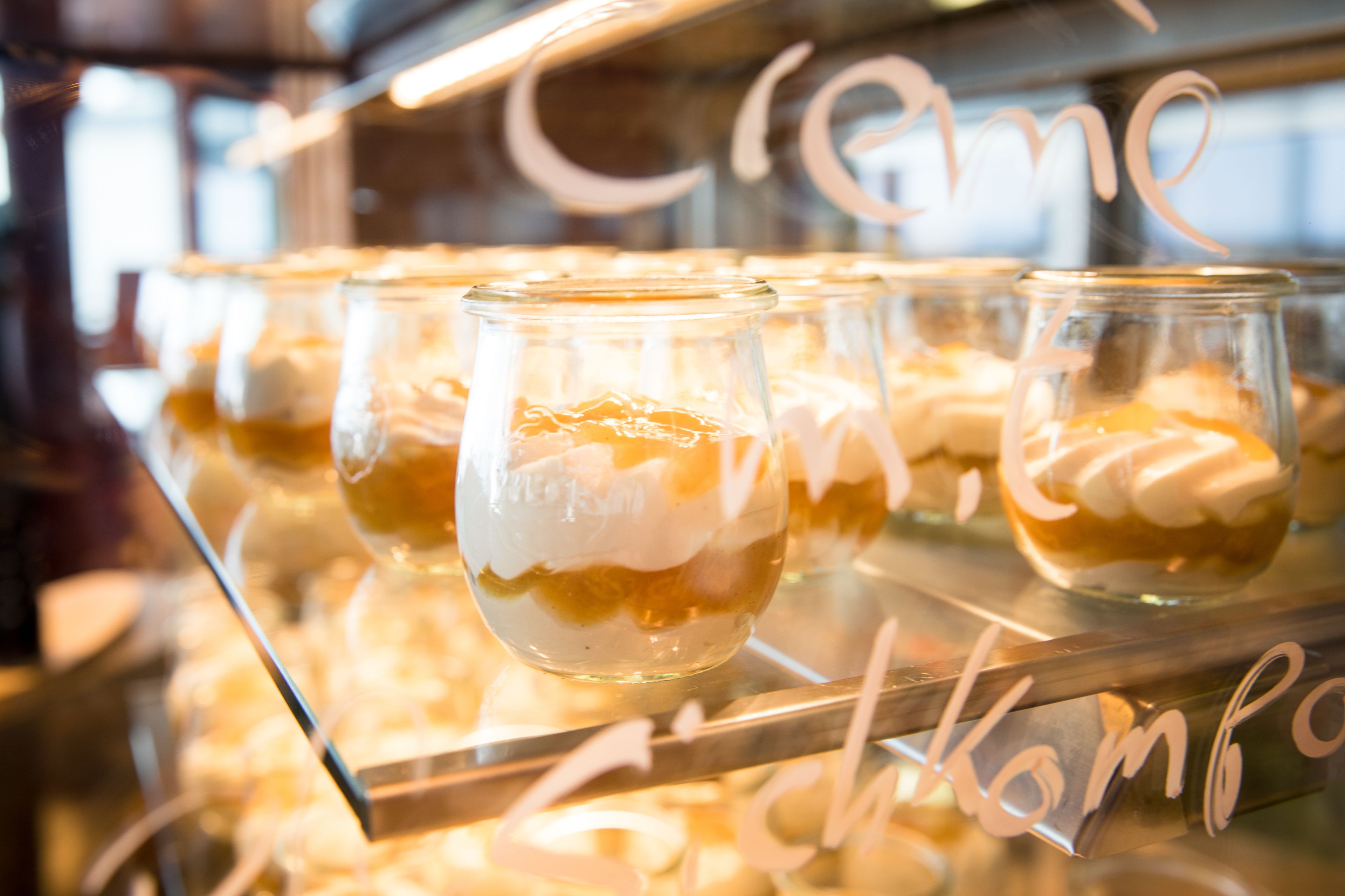 A display of desserts in clear glasses with whipped cream, arranged on a reflective surface with a sign reading "Café".
