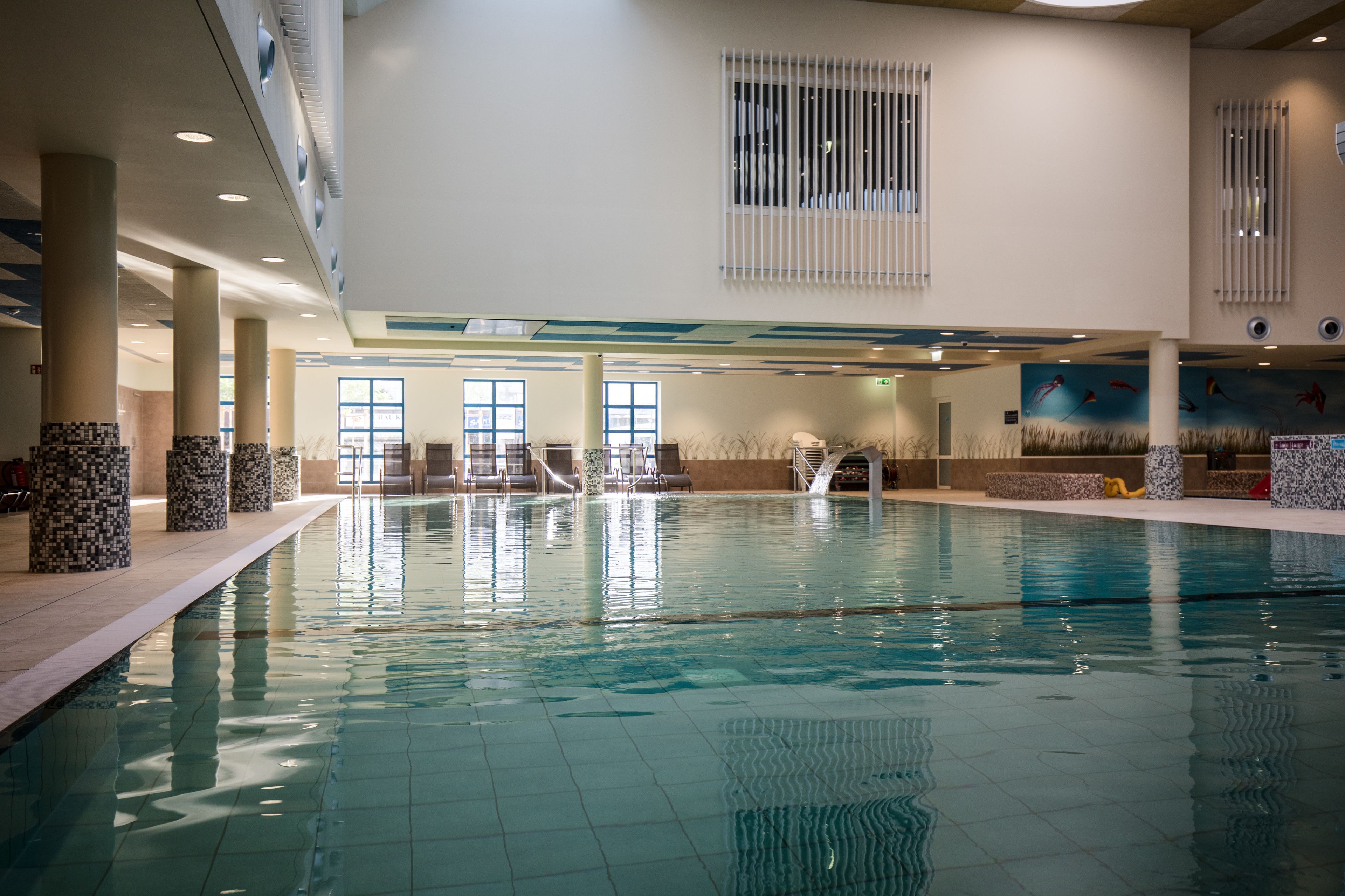En indendørs swimmingpool med klart vand, fliserede vægge og et loft med indbygget belysning.