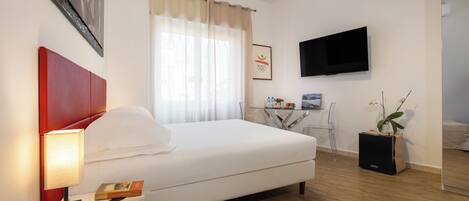 A modern hotel room with a large bed, a bedside table with a lamp, a TV mounted on the wall, and a small table with a plant.