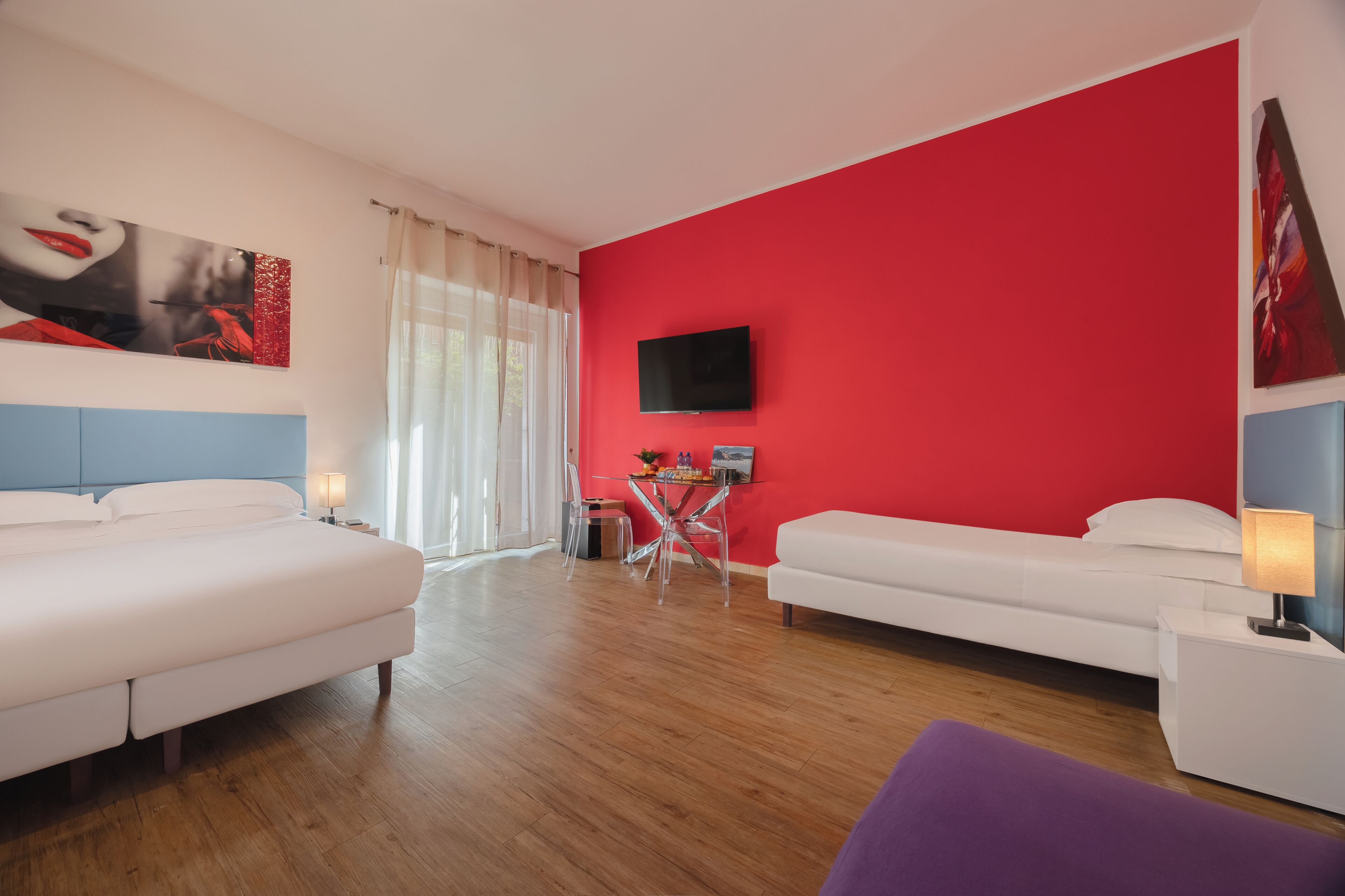 A hotel room with two beds, a red accent wall, a flat-screen TV, and a small table with chairs.