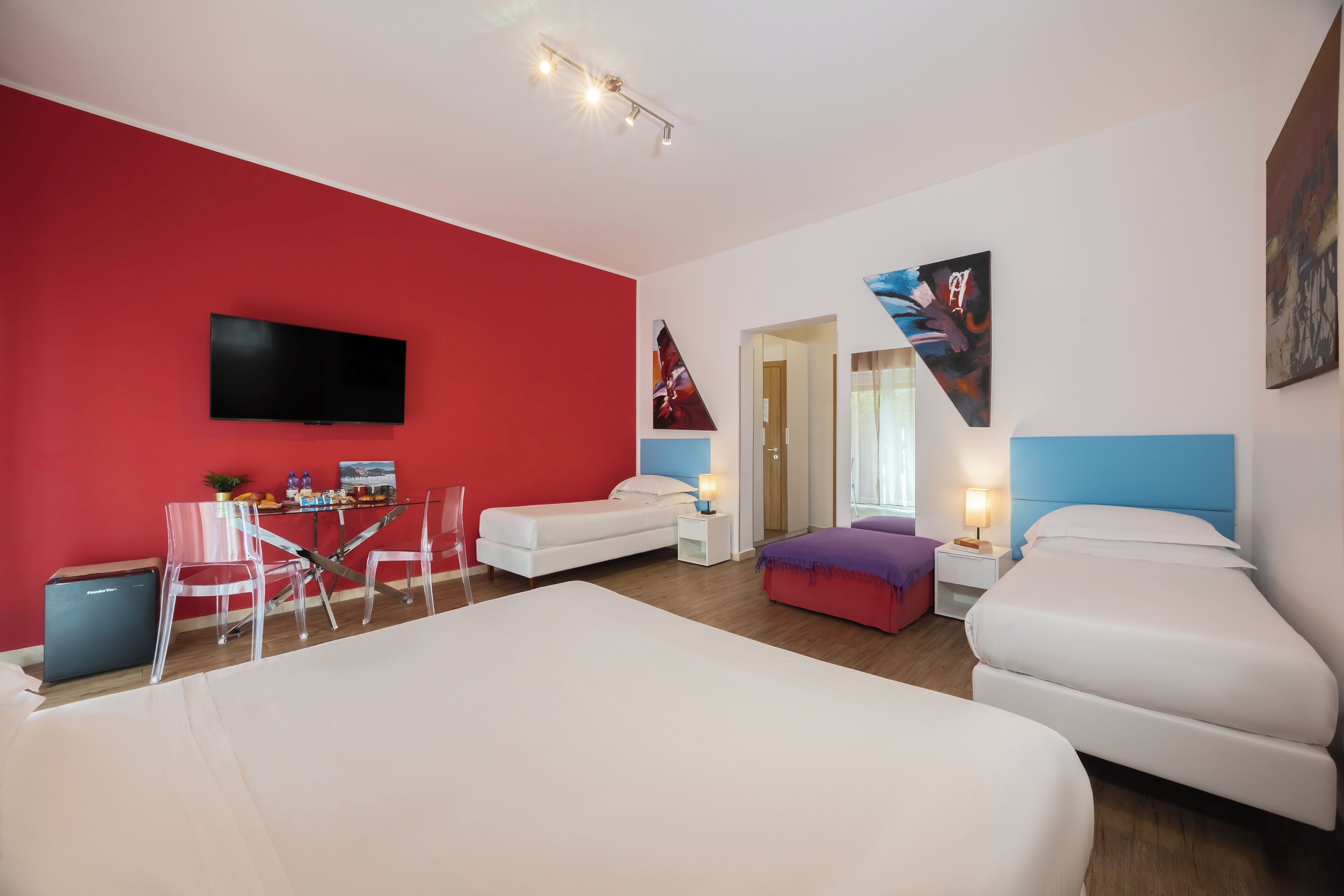A modern hotel room with a red accent wall, a flat-screen TV, a bed, a desk with a chair, and a small table with a lamp.