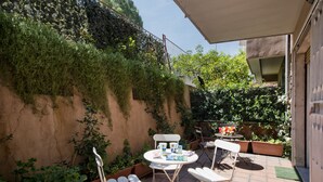 A patio with a round table, two white chairs, and a small table with a vase of flowers. - Vaticano84 (Rome)