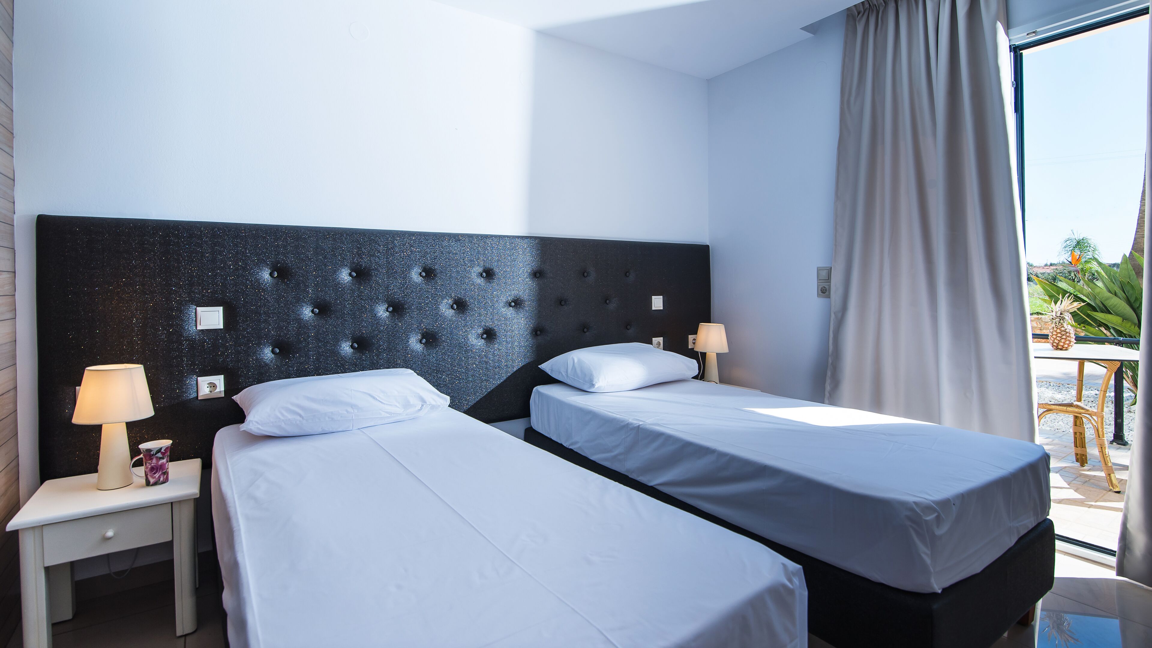 A hotel room with two beds, a nightstand, a lamp, and a view of the outdoors.
