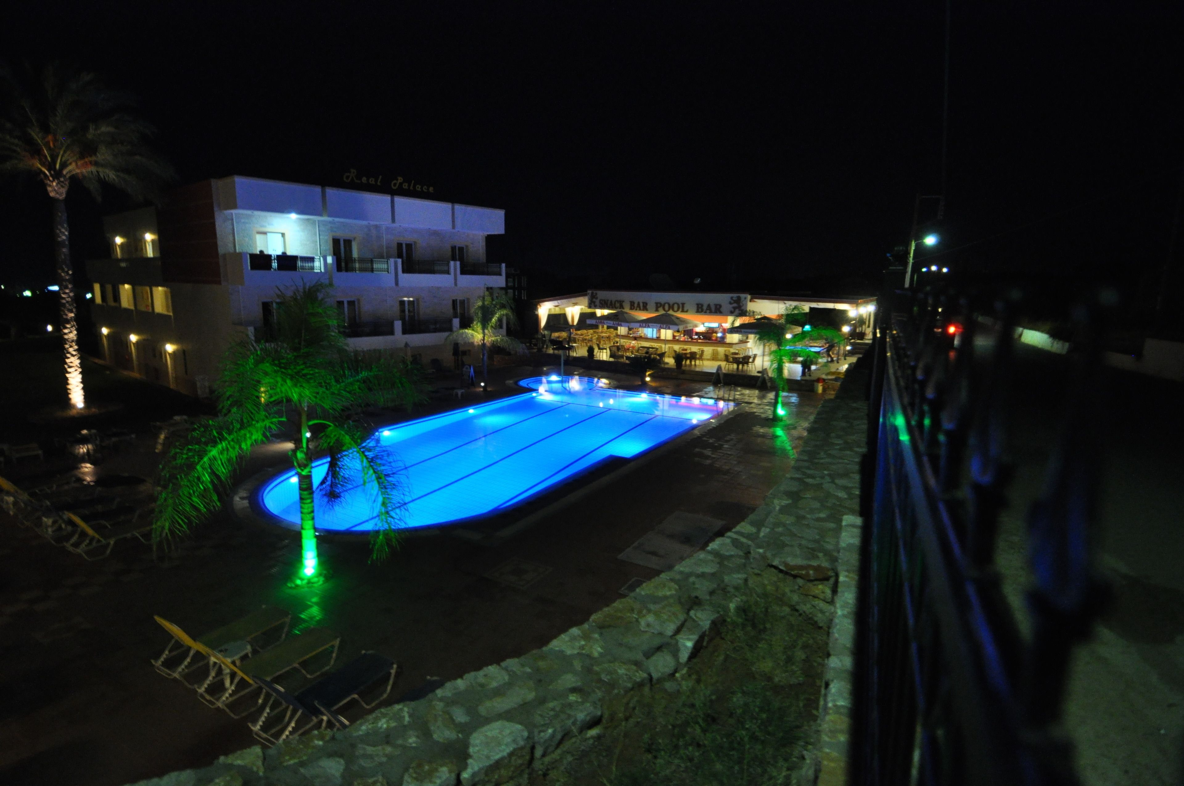 A swimming pool with a stone border, surrounded by palm trees and a building with a sign that reads "Real Palaces".