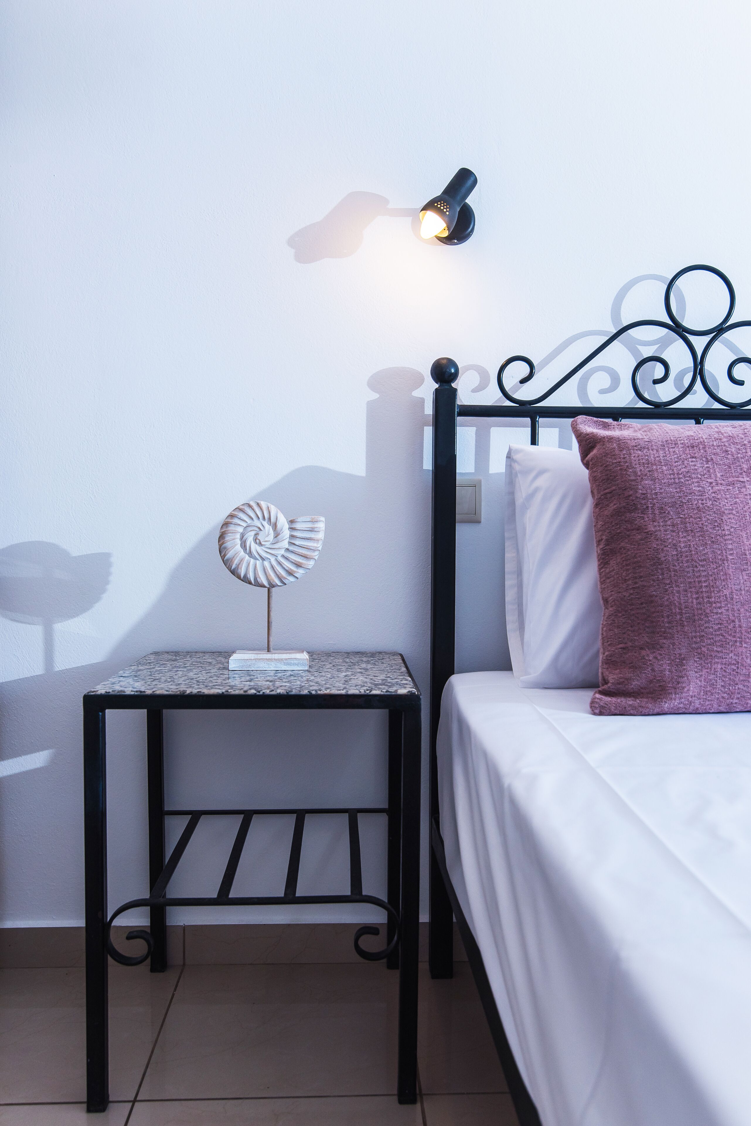 A bed with white bedding and a purple pillow, a small table with a decorative object, and a wall-mounted light fixture.