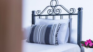 Una cama con cabecera de metal, una colcha blanca y un cojín a rayas. Hay una mesita de noche pequeña con una lámpara blanca y una planta en maceta con flores rosas.