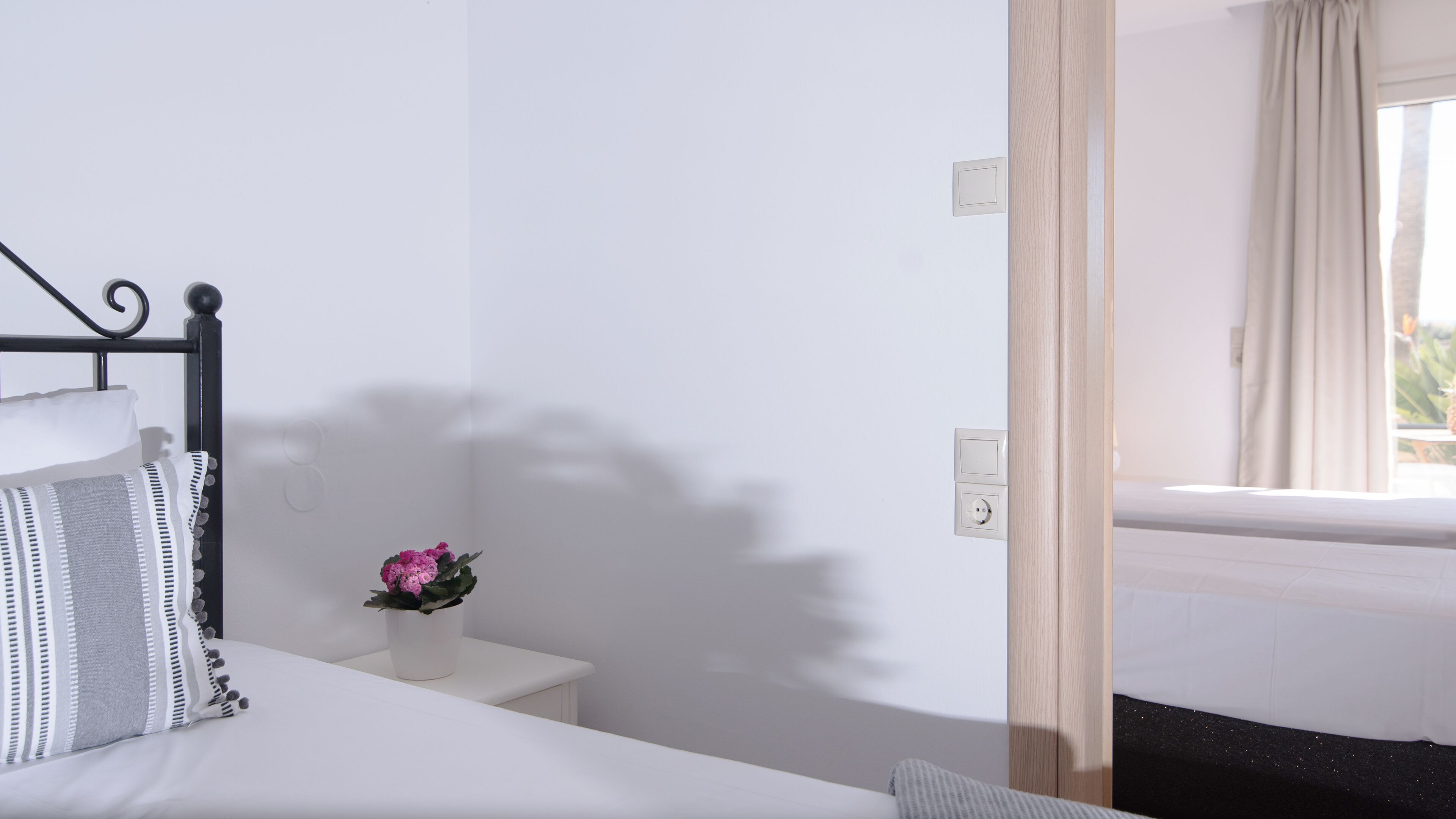 A bedroom with a bed, a bedside table with a flower, a wall socket, and a window with curtains.