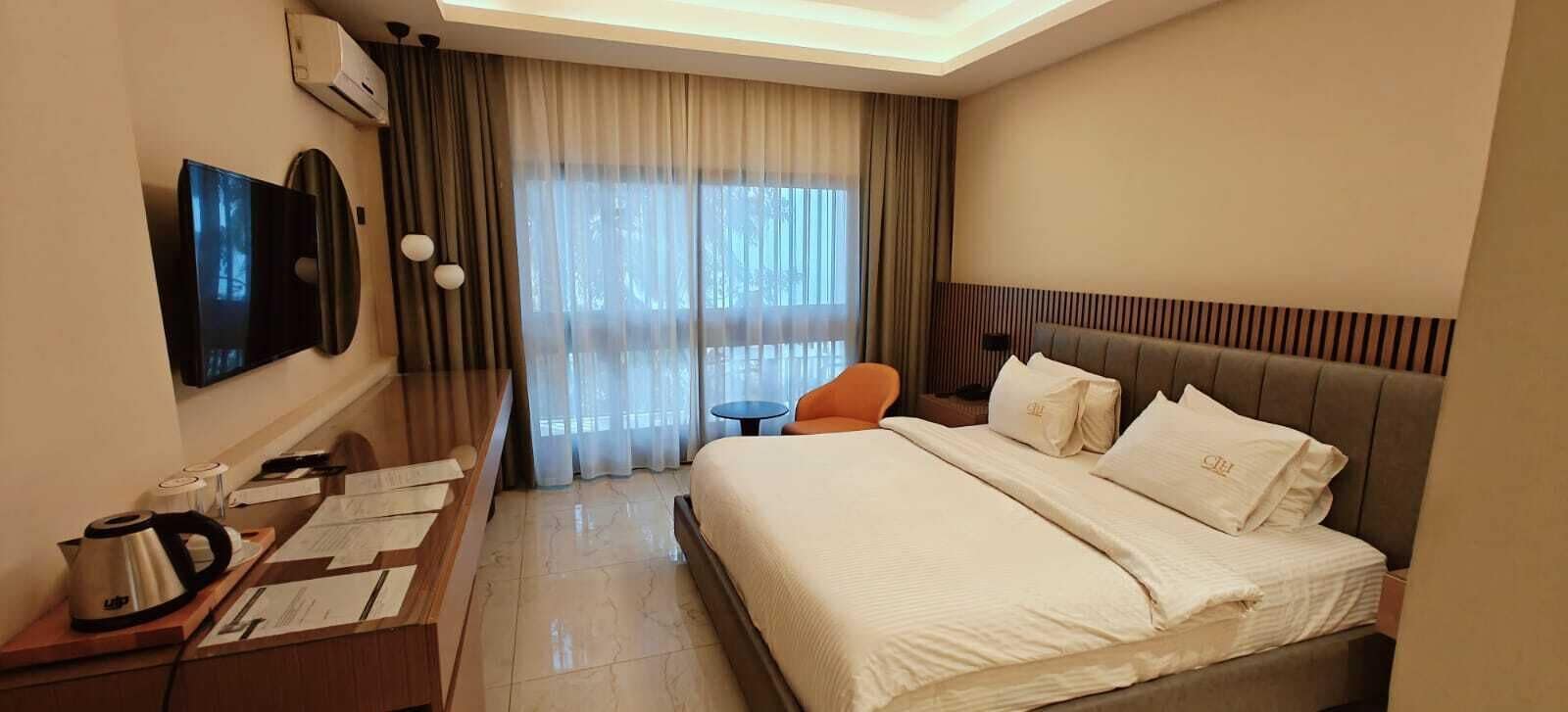 A hotel room with a bed, a desk with a kettle, a chair, a TV, and a large window with curtains.