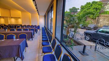 A conference room with rows of tables and chairs, large windows, and a view of parked cars and greenery outside.