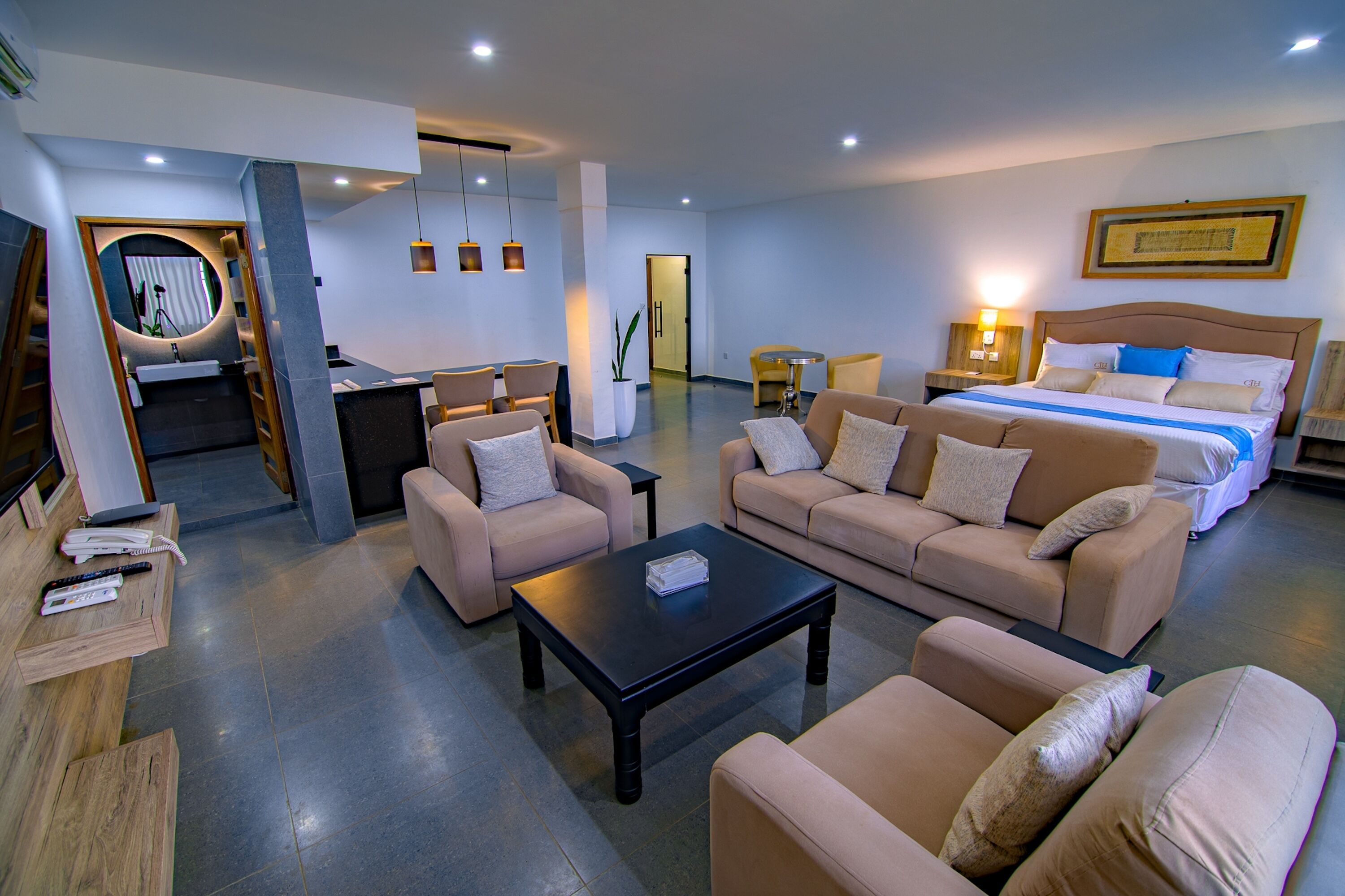A modern hotel room with a large bed, two sofas, a coffee table, and a dining area with a small table and chairs.