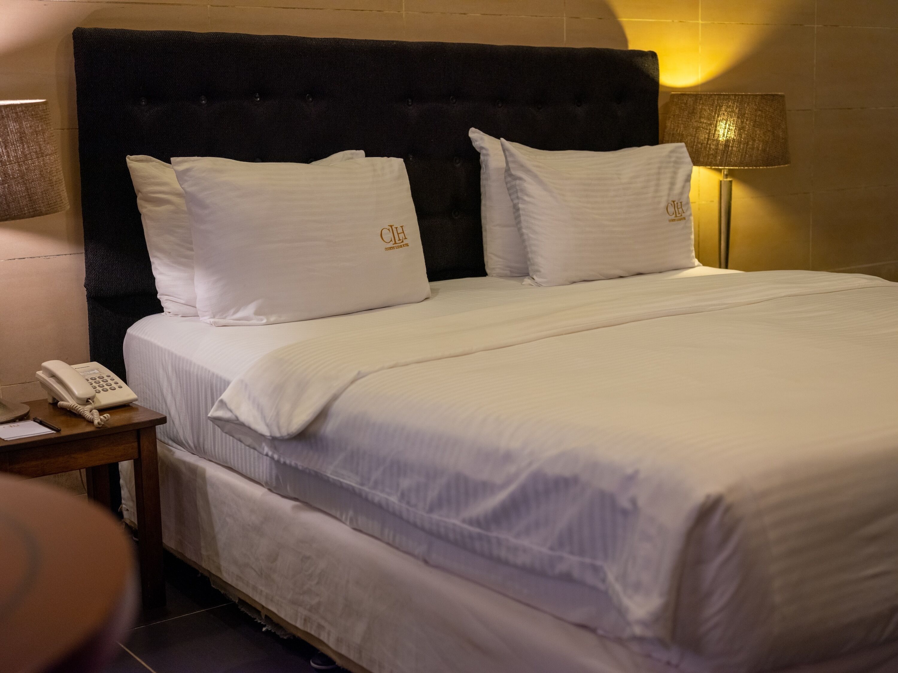 Una habitación de hotel con una cama grande, ropa de cama blanca, cabecera oscura y teléfono en la mesita de noche.