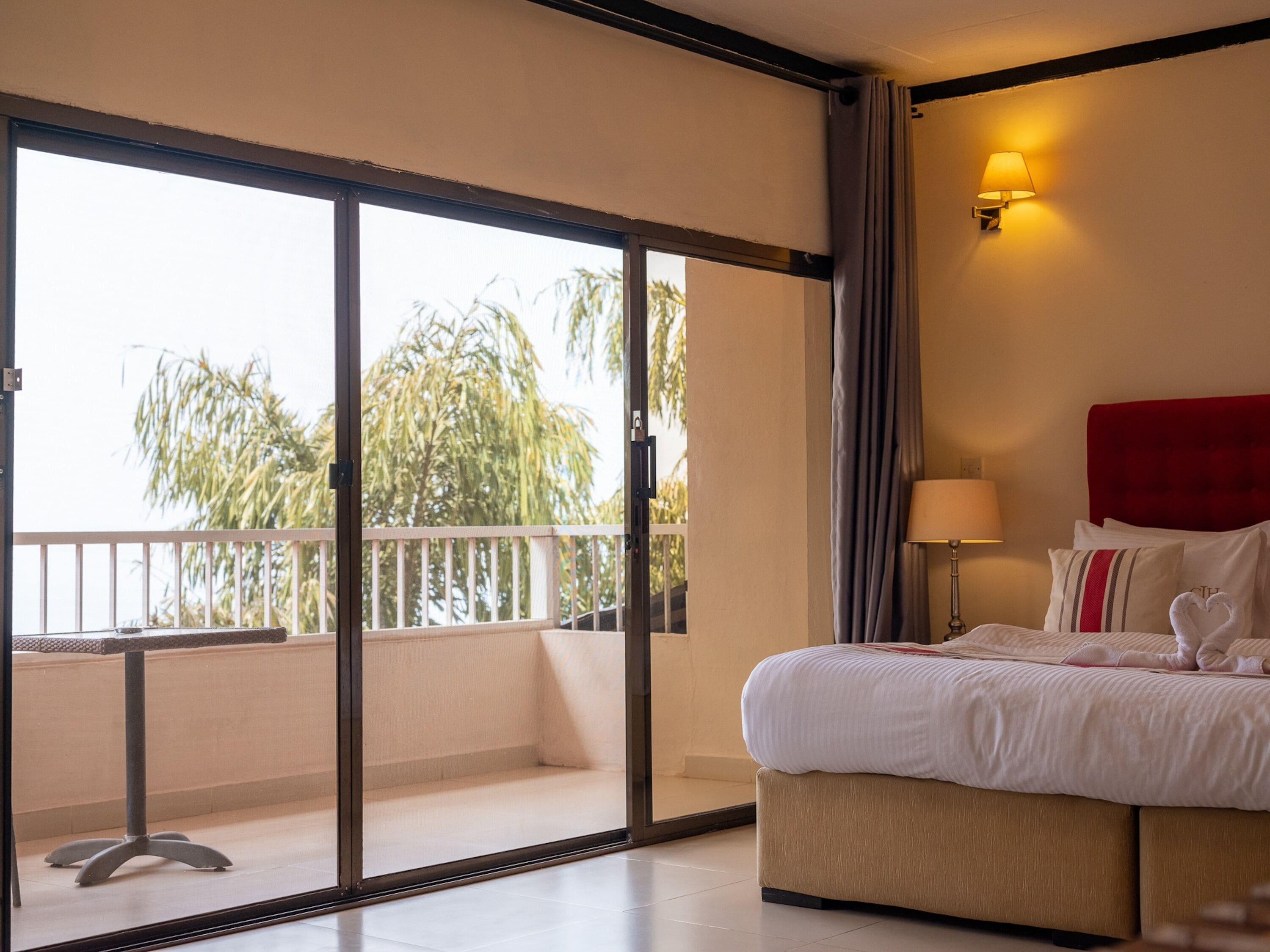 A hotel room with a bed, a nightstand, a lamp, a sliding glass door to a balcony, and a view of palm trees.