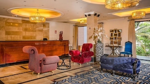 A reception area with a wooden reception desk, red and blue upholstered chairs, and a large vase with white flowers. - The Country Lodge Hotel (Freetown)