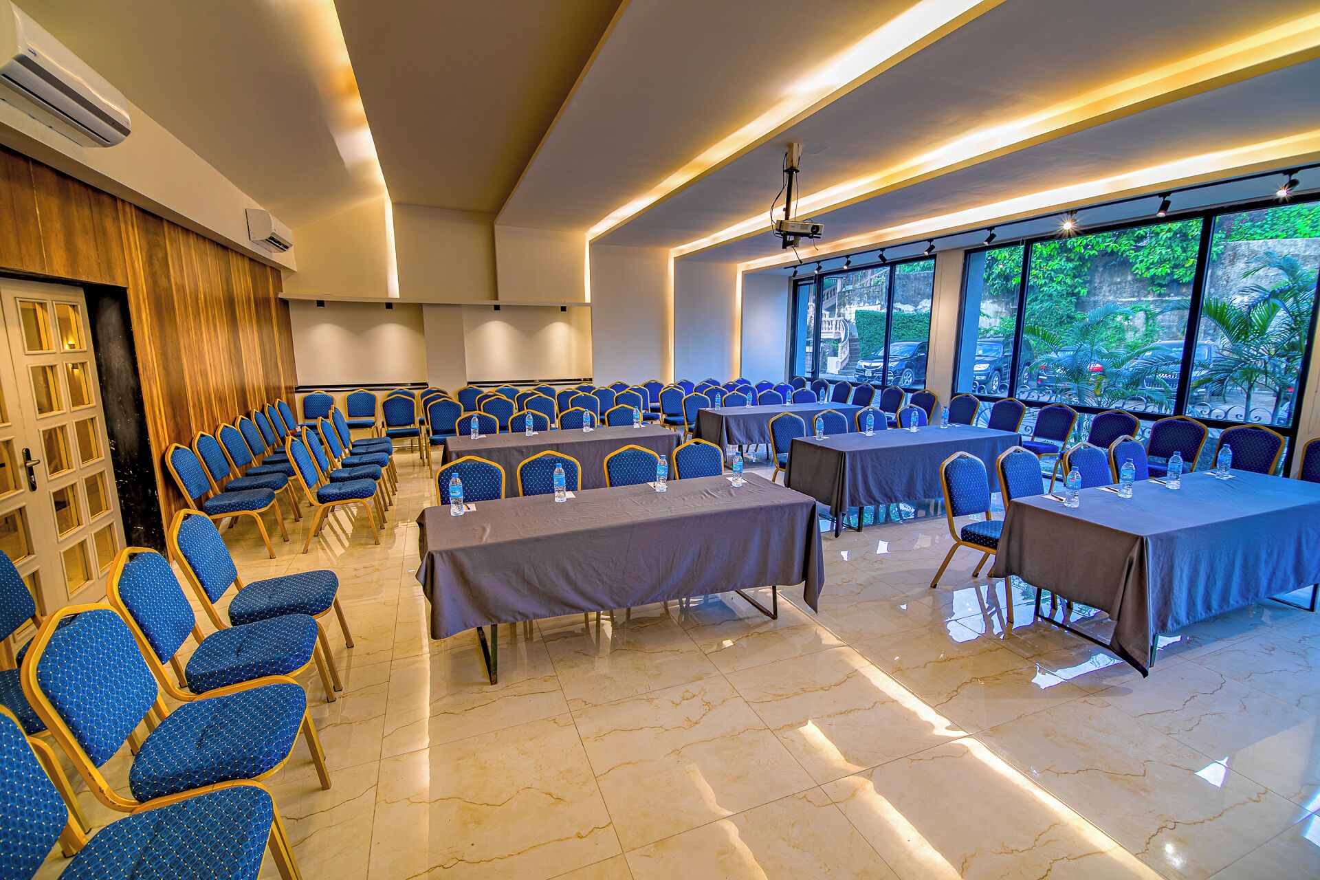 Ein Konferenzraum mit blauen Stühlen und Tischen, großen Fenstern mit Blick auf Grünflächen und modernen Leuchten.