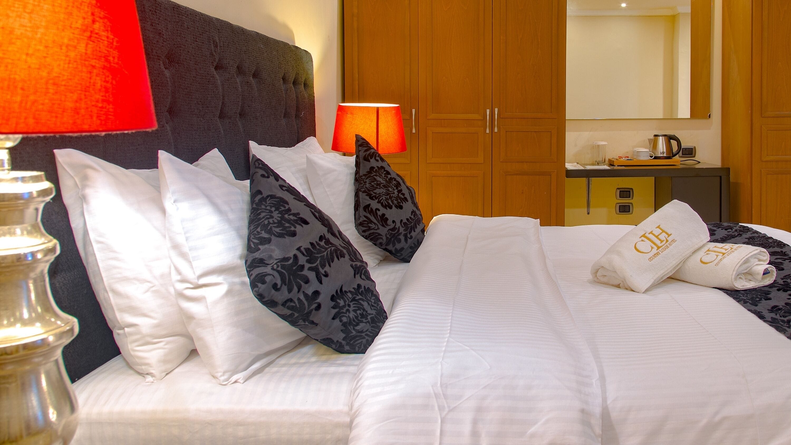 Una cama bien hecha con sábanas blancas y almohadas decorativas, una lámpara de noche y un armario de madera al fondo.