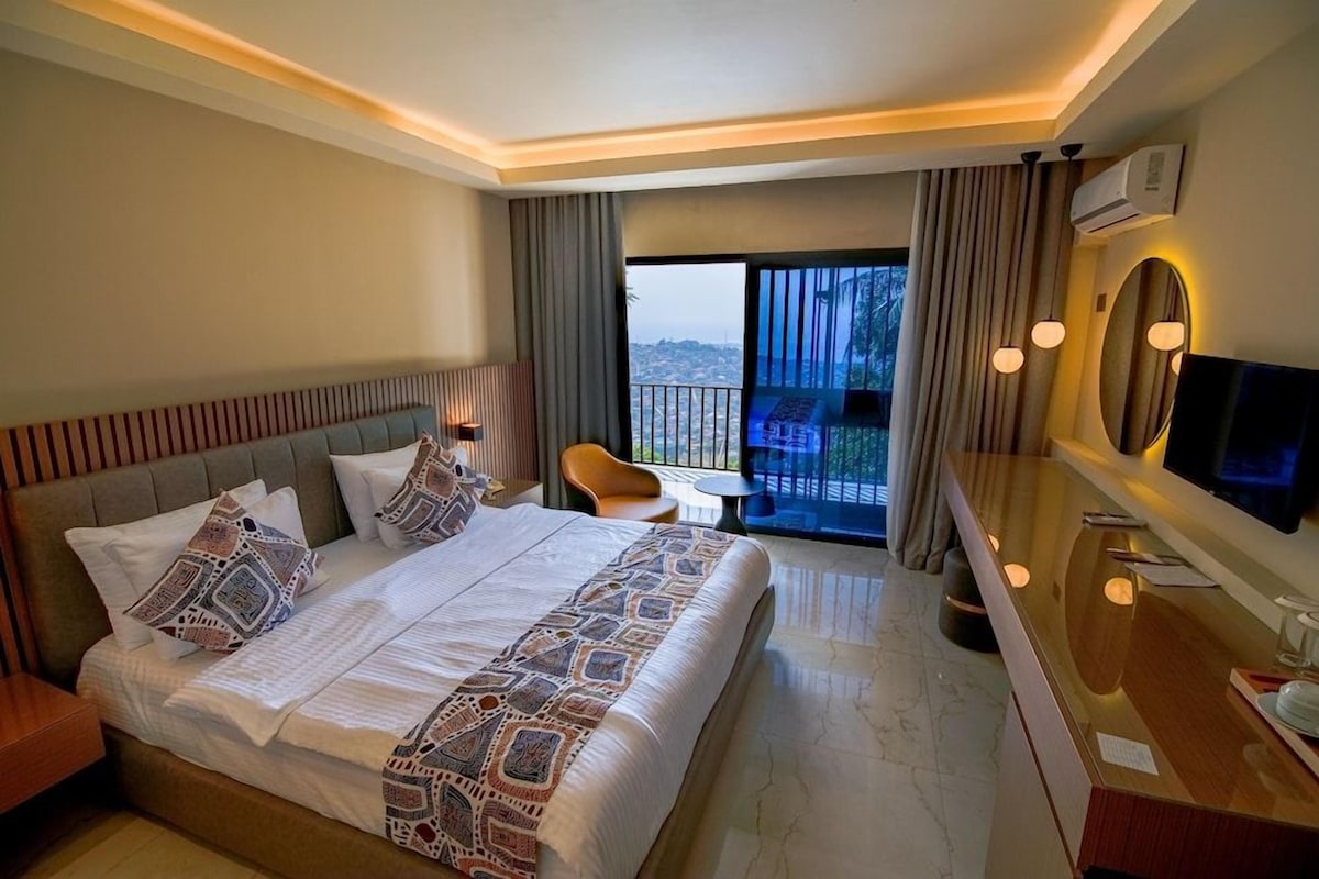 A hotel room with a large bed, a desk, a chair, a TV, and a balcony with a city view.