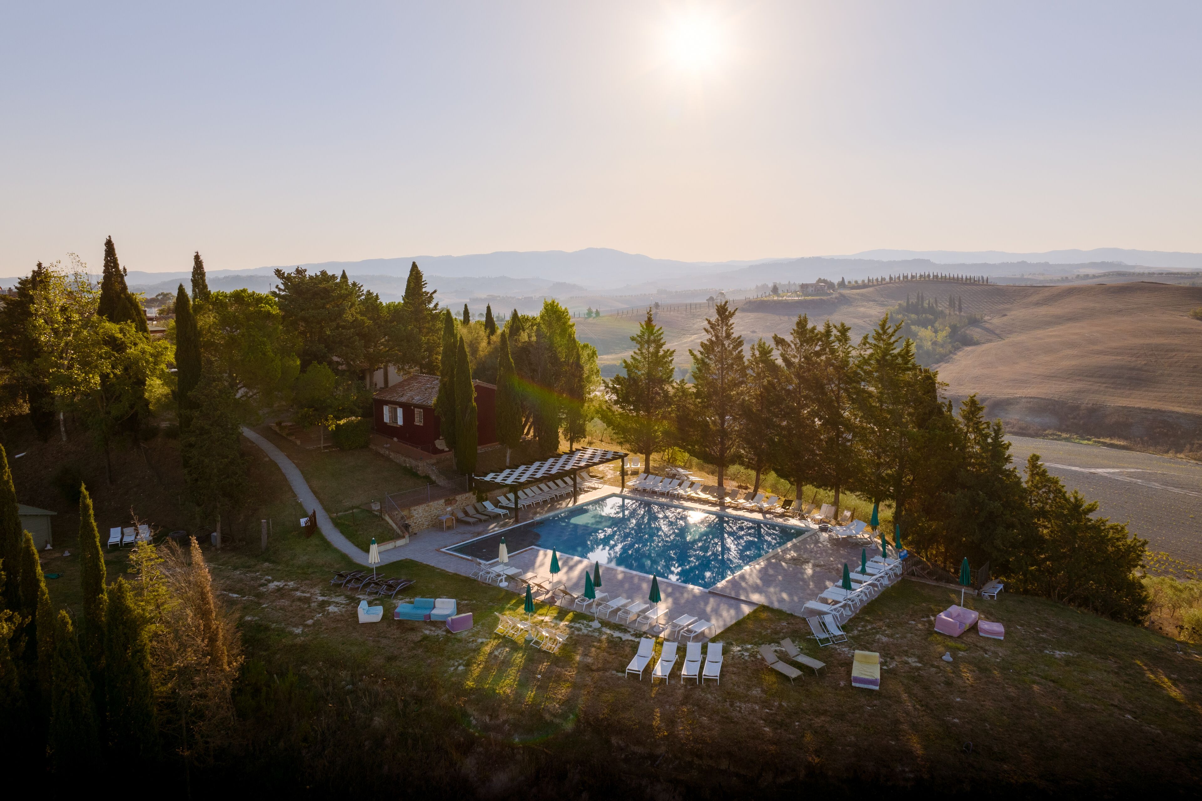 Un área de piscina con sillones de descanso y sombrillas, rodeada de árboles y colinas al fondo.