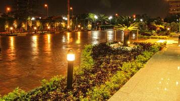 A nighttime view of a waterfront promenade with a paved walkway, illuminated street lamps, and landscaped greenery.