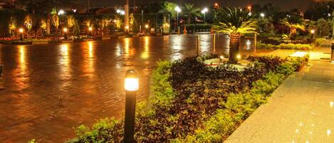 A nighttime view of a waterfront promenade with a paved walkway, illuminated street lamps, and landscaped greenery.