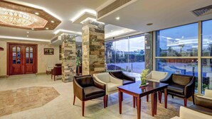 A spacious lobby with a stone column, leather chairs, a wooden table, and a large window offering a view of the outdoors. - Mirita Hotel (Tenth of Ramadan City)