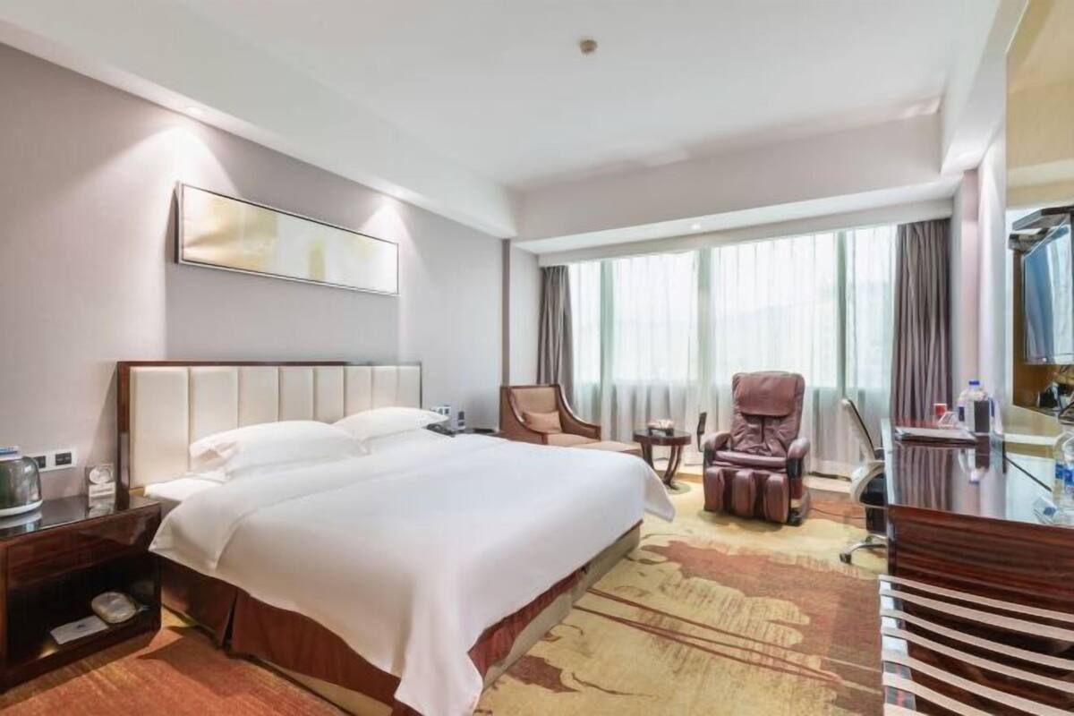 Ein Hotelzimmer mit einem großen Bett, einem Massagesessel, einem Schreibtisch und einem großen Fenster mit Vorhängen.