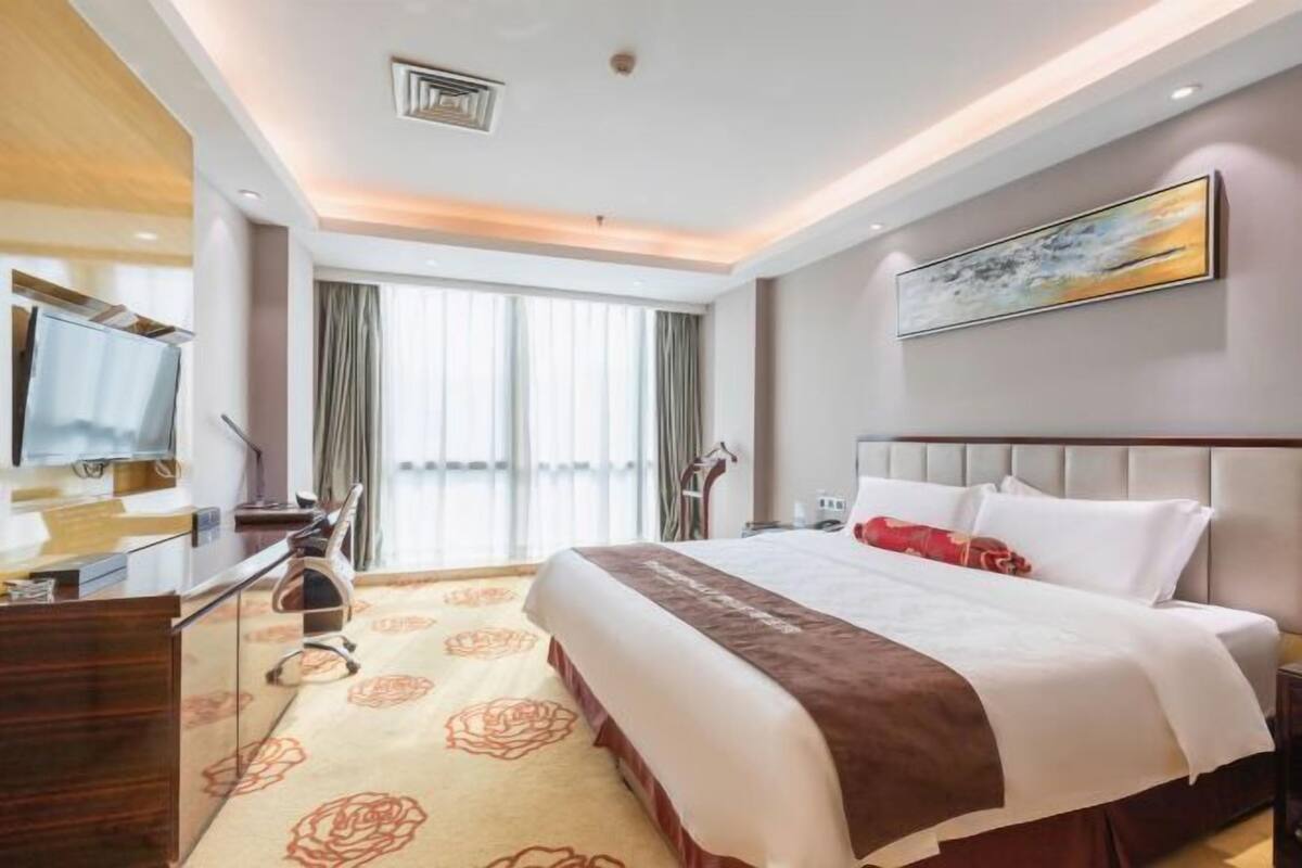 Ein Hotelzimmer mit einem großen Bett, einem Schreibtisch mit Stuhl, einem Fernseher und einem Fenster mit Vorhängen.