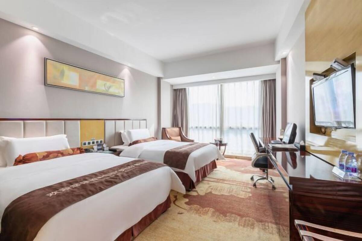 Ein Hotelzimmer mit zwei Betten, einem Schreibtisch, einem Stuhl, einem Fernseher und einem großen Fenster mit Vorhängen.