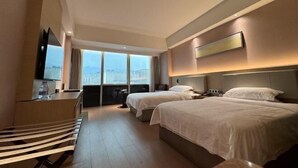 Habitación de hotel con dos camas, un escritorio, un televisor y un amplio ventanal con vista a la ciudad.