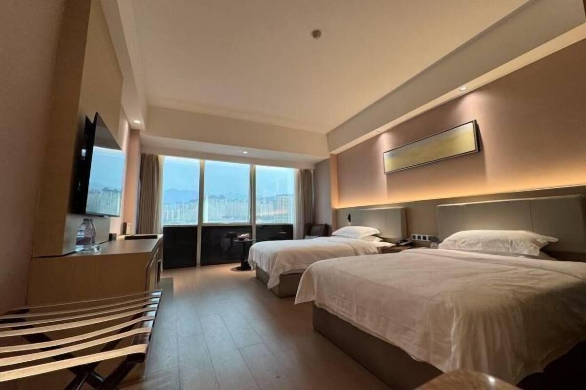 Ein Hotelzimmer mit zwei Betten, einem Schreibtisch, einem Fernseher und einem großen Fenster mit Blick auf die Stadt.