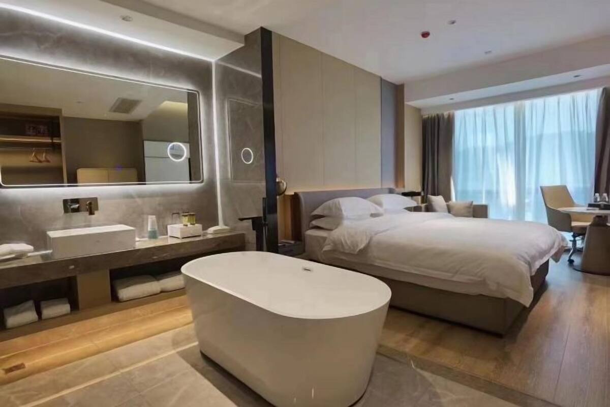 Ein modernes Hotelzimmer mit einem großen Bett, einem separaten Badezimmer mit Badewanne, einem großen Spiegel und Blick ins Freie.