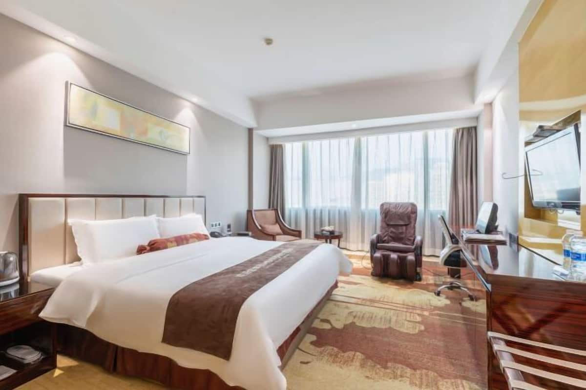 Ein Hotelzimmer mit einem großen Bett, einem Schreibtisch mit Stuhl, einem Fernseher und einem Fenster mit Vorhängen.