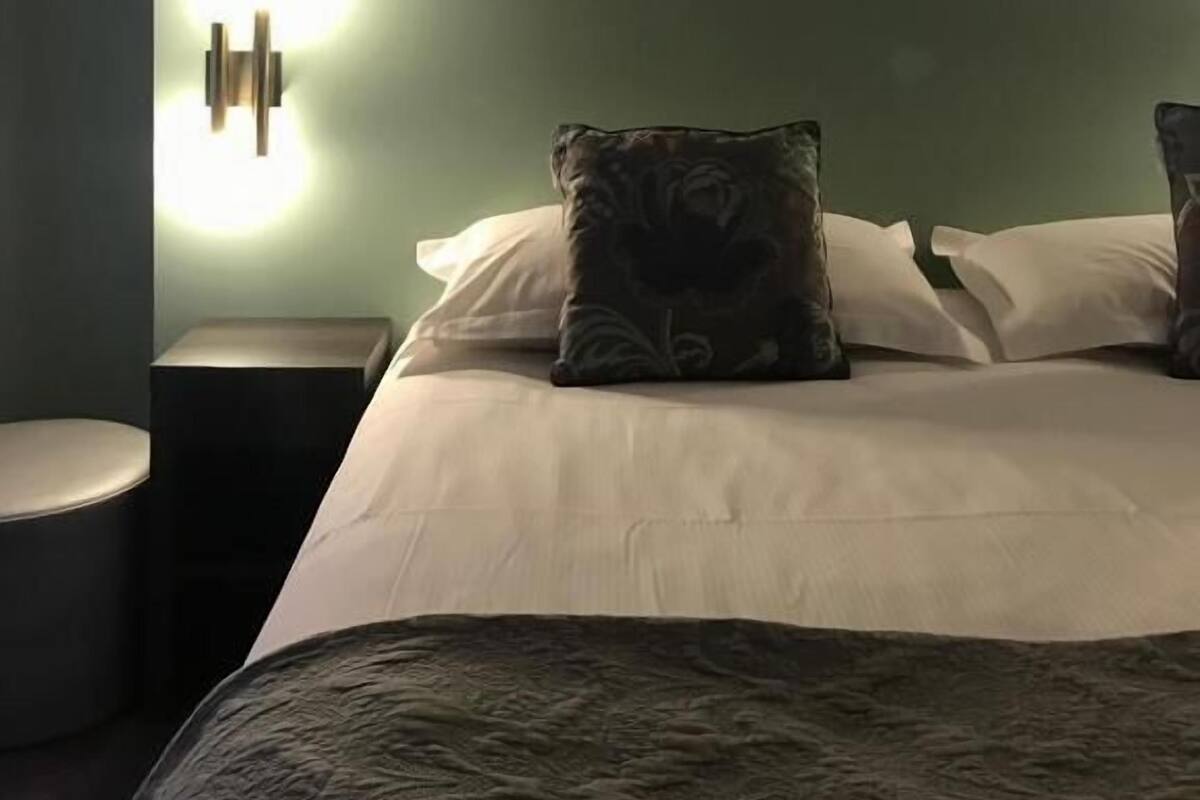 A bedroom with a bed, two pillows, a framed picture on the wall, and a wall-mounted light fixture.