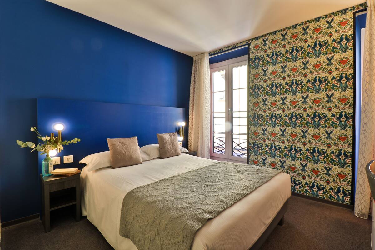 A bedroom with a large bed, a nightstand with a lamp, and a window with patterned curtains.