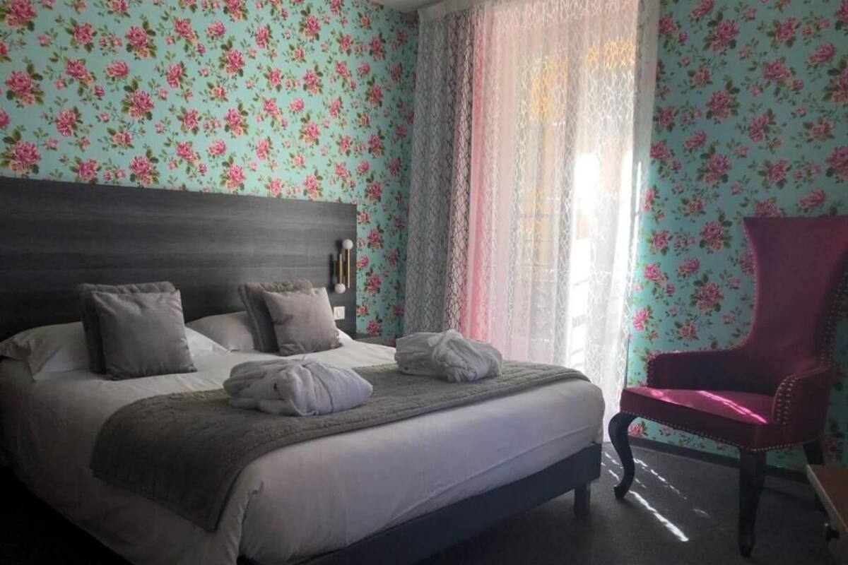 A neatly made bed with pillows and a blanket, a red chair, and floral wallpaper.