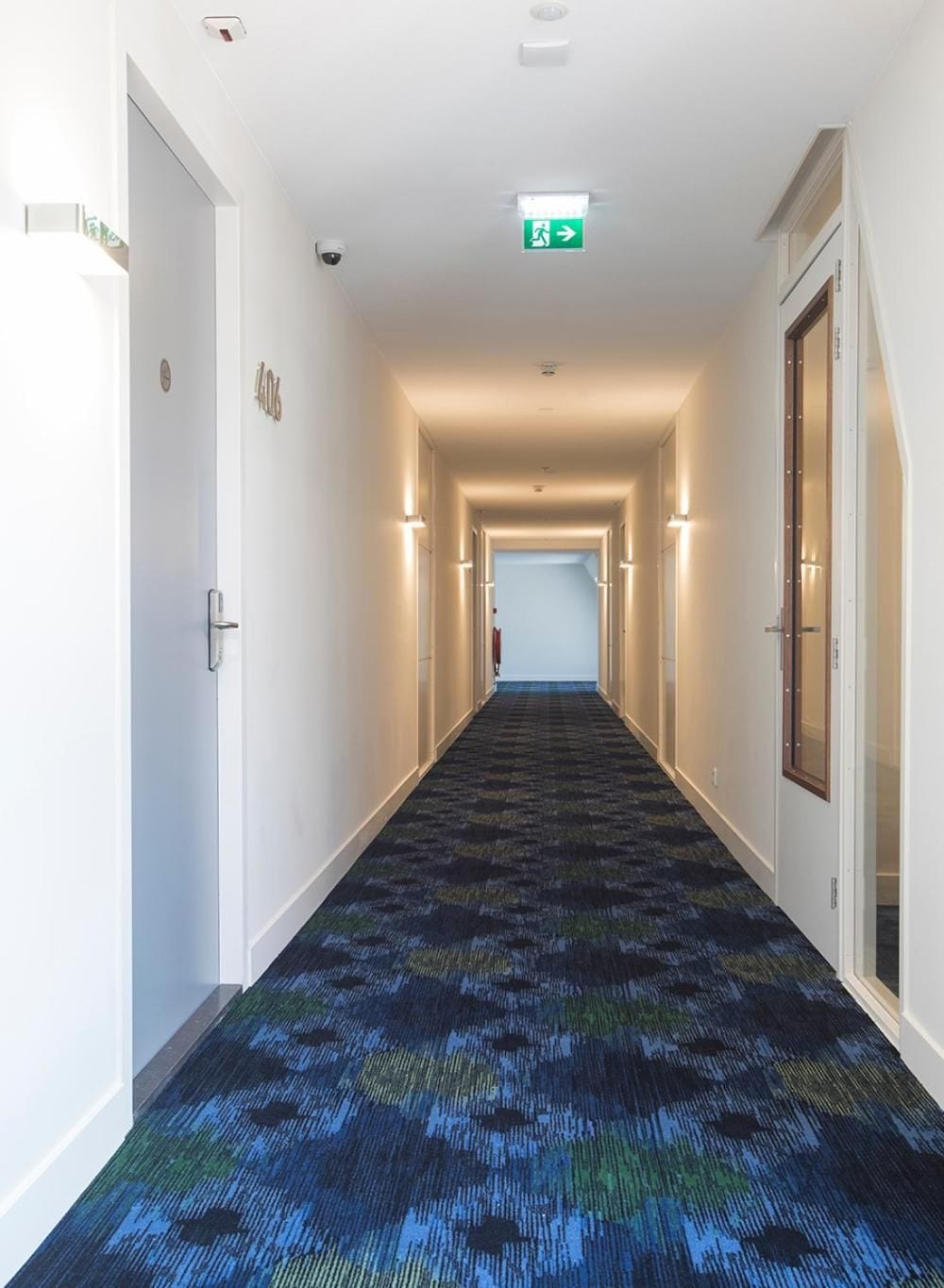 Un corridoio di un hotel con pavimenti rivestiti di moquette, porte su entrambi i lati e un segnale di uscita verde sul soffitto.