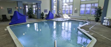 An indoor swimming pool with clear water, a diving board, and a seating area with chairs and potted plants.