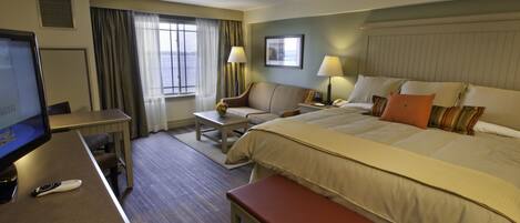 A hotel room with a bed, a desk with a computer, a chair, a small table with a lamp, and a window with curtains.