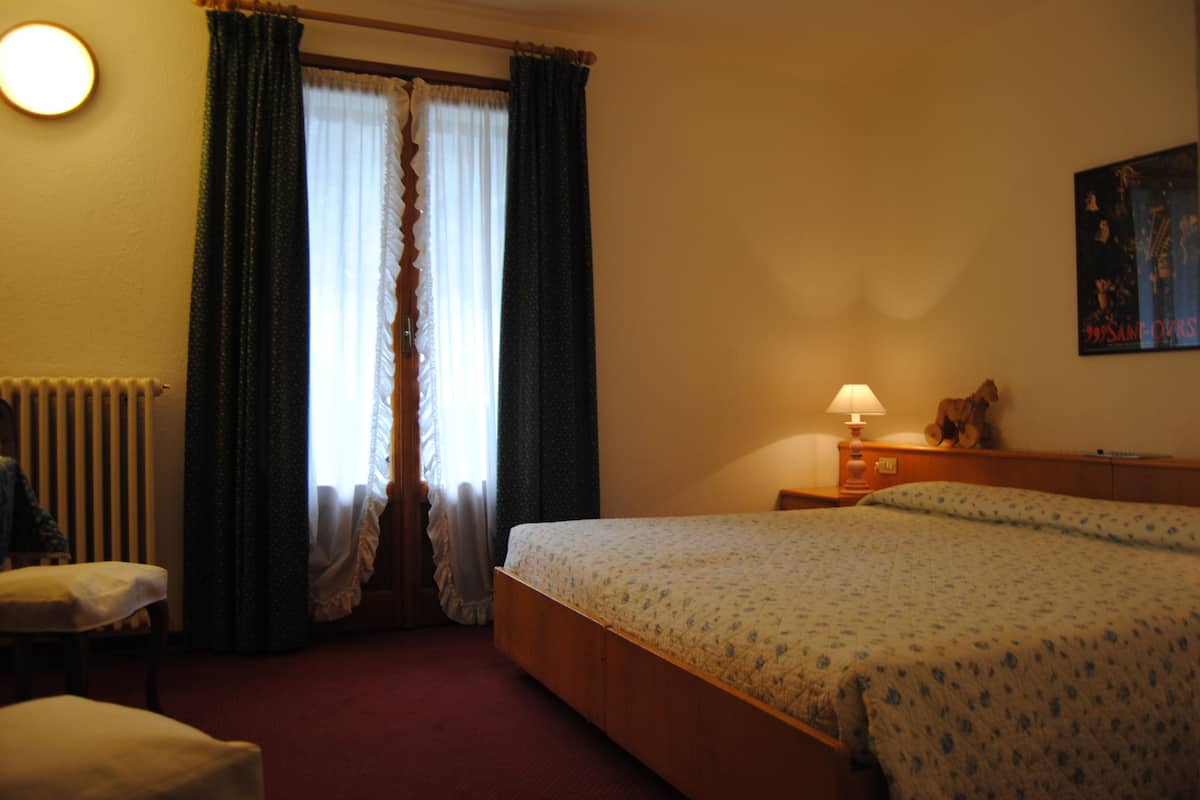 A hotel room with a bed, a nightstand, a lamp, a radiator, and a window with curtains.