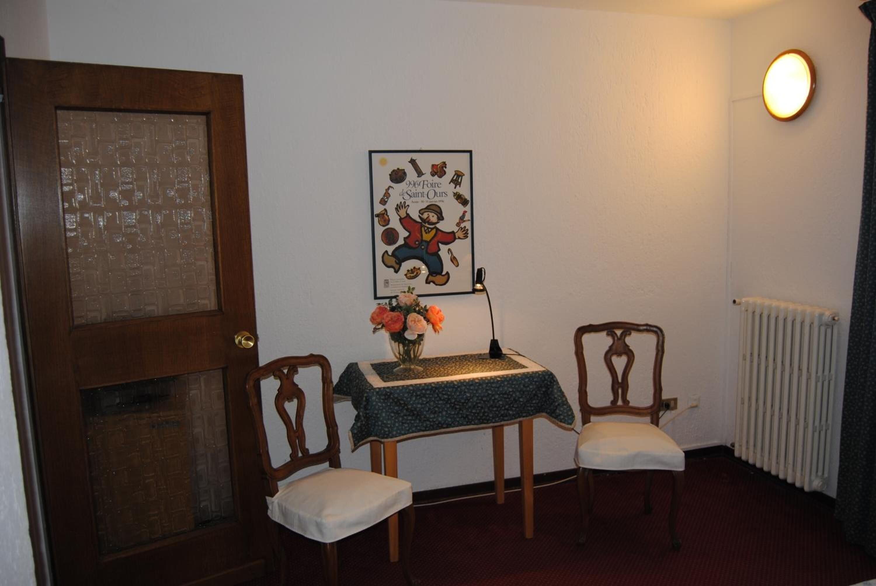 Una habitación con una puerta de madera, un cartel enmarcado en la pared, una mesita con un jarrón de flores, dos sillas de madera y un radiador.