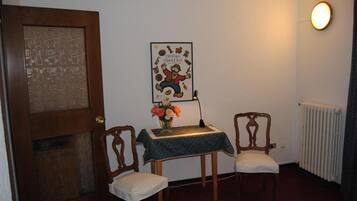 Una habitación con una puerta de madera, un cartel enmarcado en la pared, una mesita con un jarrón de flores, dos sillas de madera y un radiador.