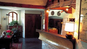 Un mostrador de recepción de madera con una lámpara, una planta en maceta y una alfombra roja en una habitación con pared de piedra y puerta de madera.