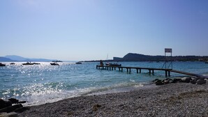 Una playa pedregosa con un muelle de madera que se adentra en aguas tranquilas, barcos amarrados y una cadena montañosa al fondo.