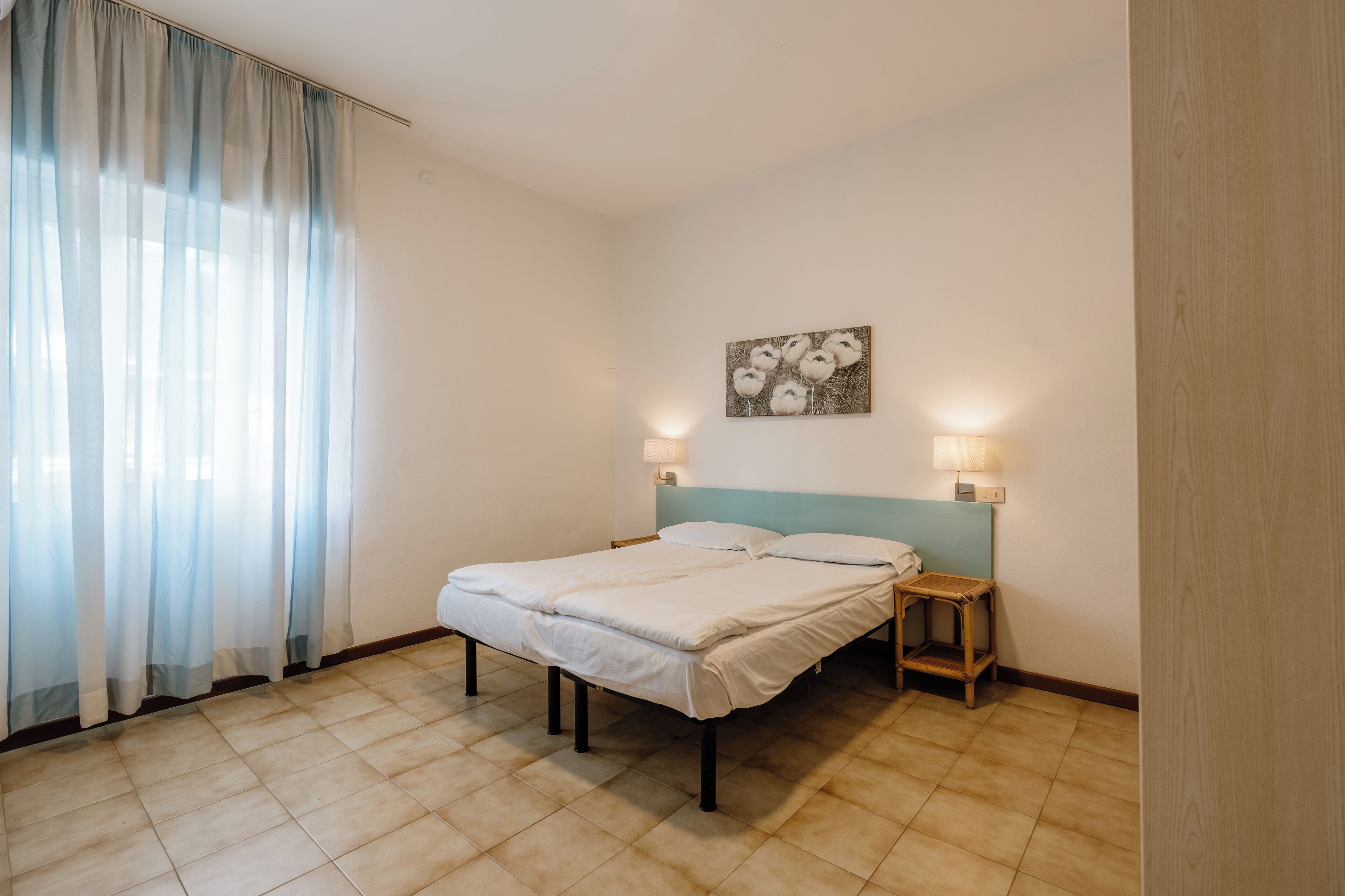 A bedroom with a bed, two bedside tables, a wall-mounted lamp, and a window with sheer curtains.