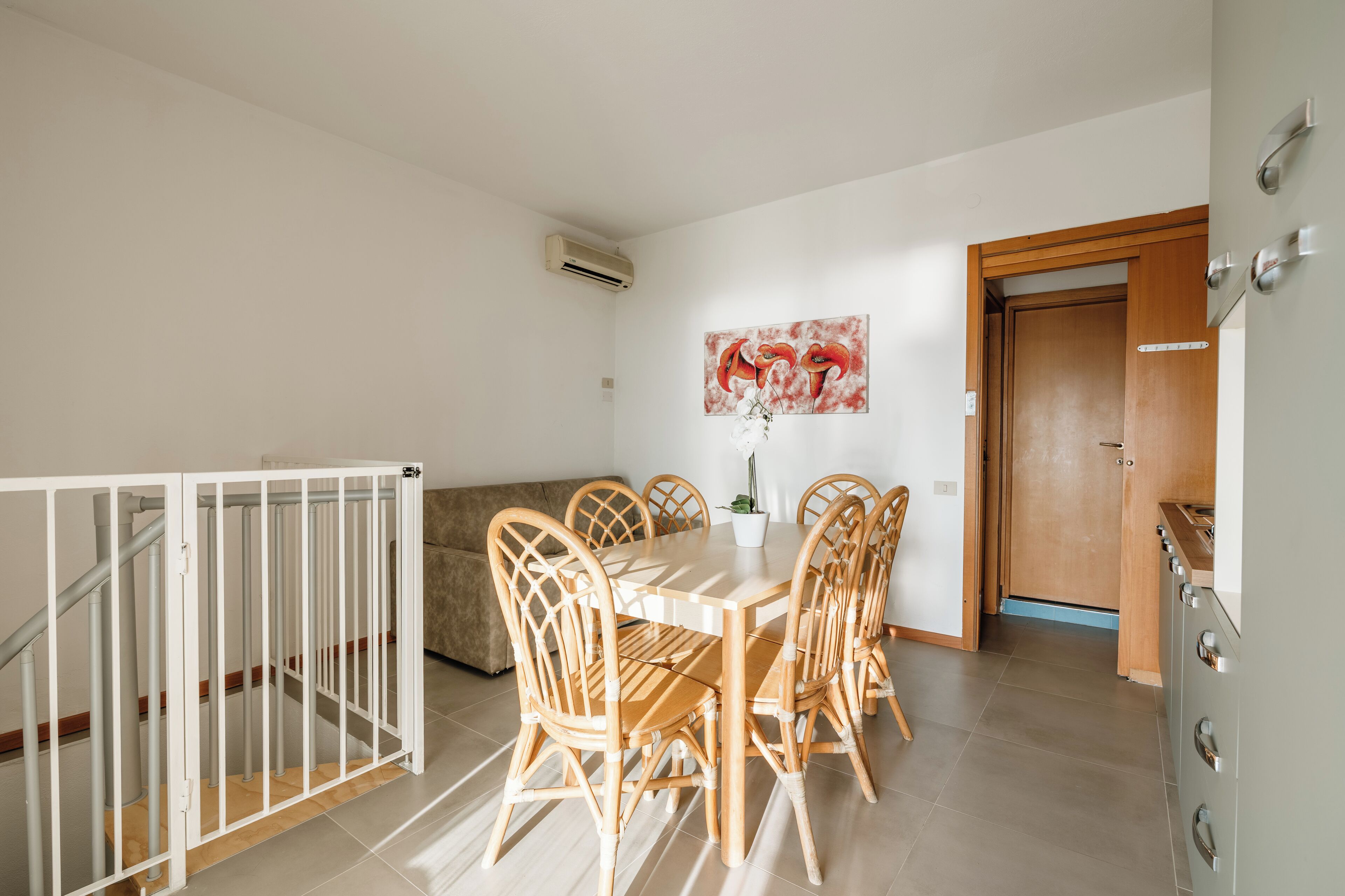 Un comedor con una mesa blanca y sillas de madera, un cuadro en la pared y una zona de cocina con armarios.