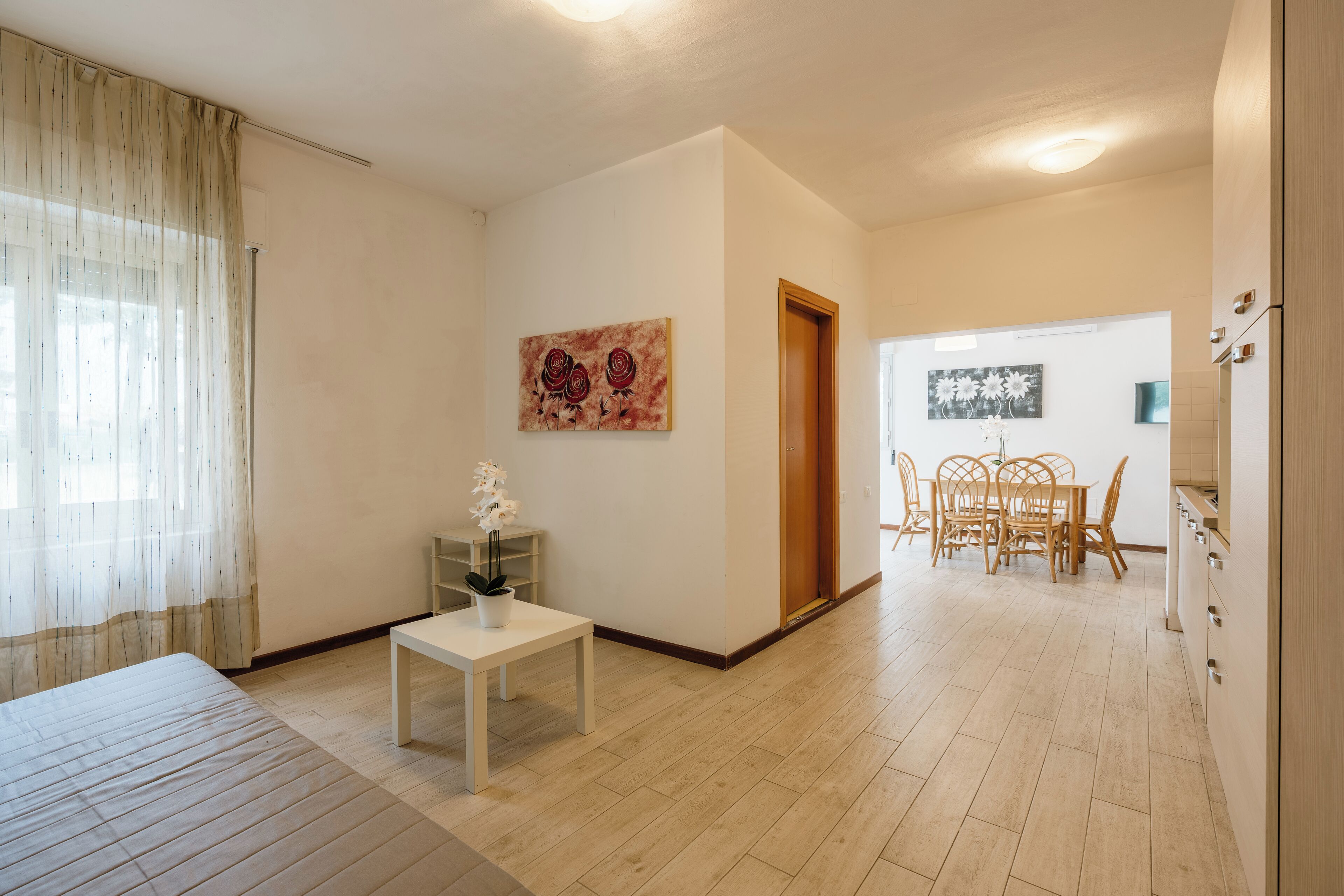 Una camera d'albergo moderna con un letto, un piccolo tavolino con una pianta in vaso, un'area ristorazione con tavolo e sedie, e un quadro appeso alla parete.