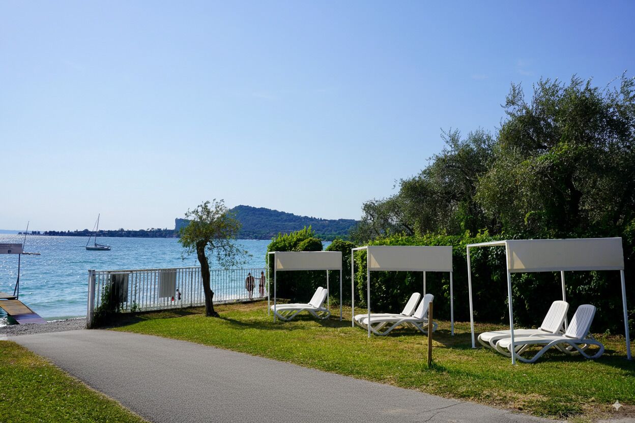 Ein Seeufer mit drei weißen Liegestühlen unter weißen Sonnensegeln, einem gepflasterten Weg und Blick auf den See mit Booten und fernen Hügeln.