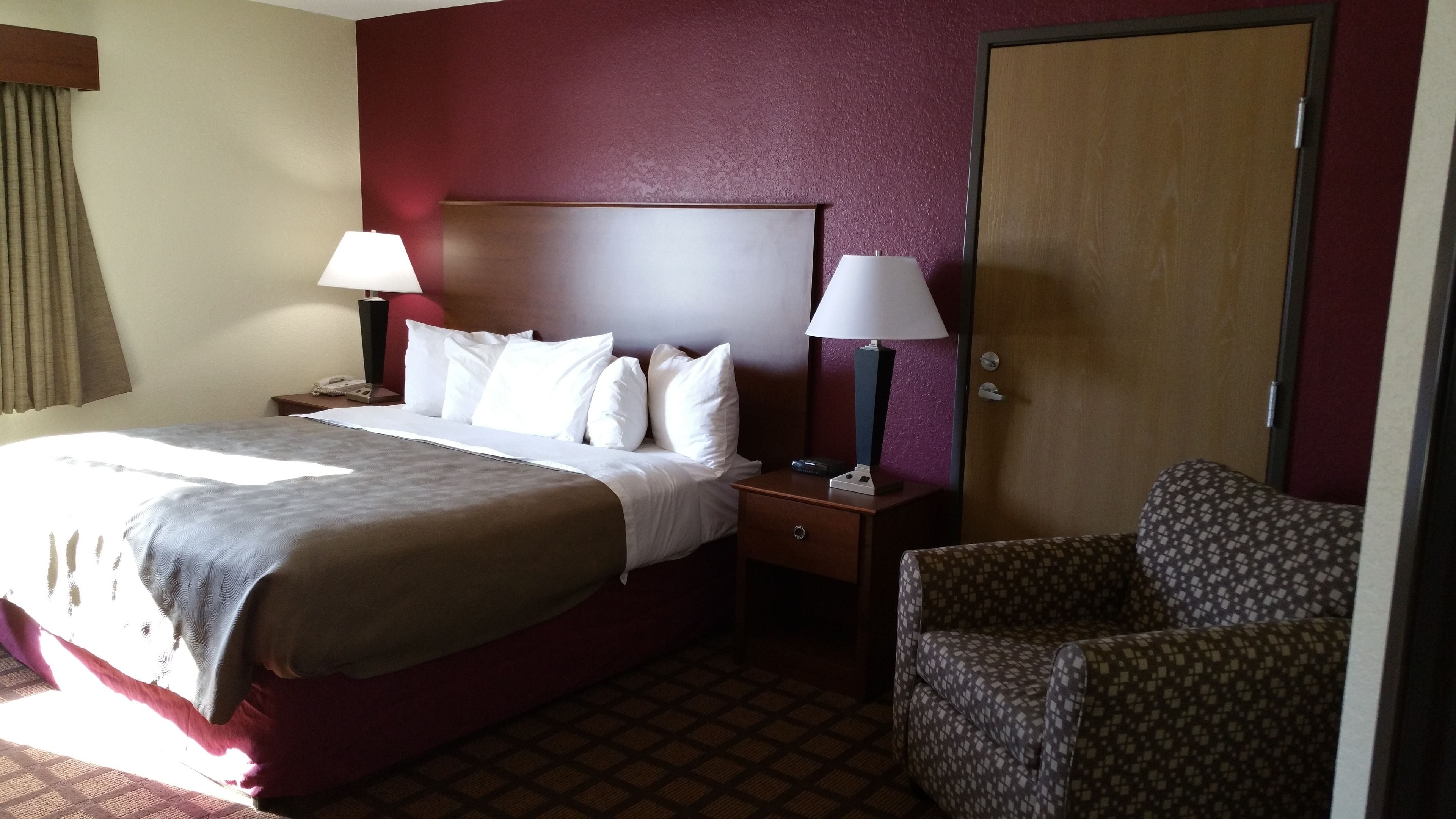 A hotel room with a large bed, two bedside tables with lamps, a patterned armchair, and a wooden door.