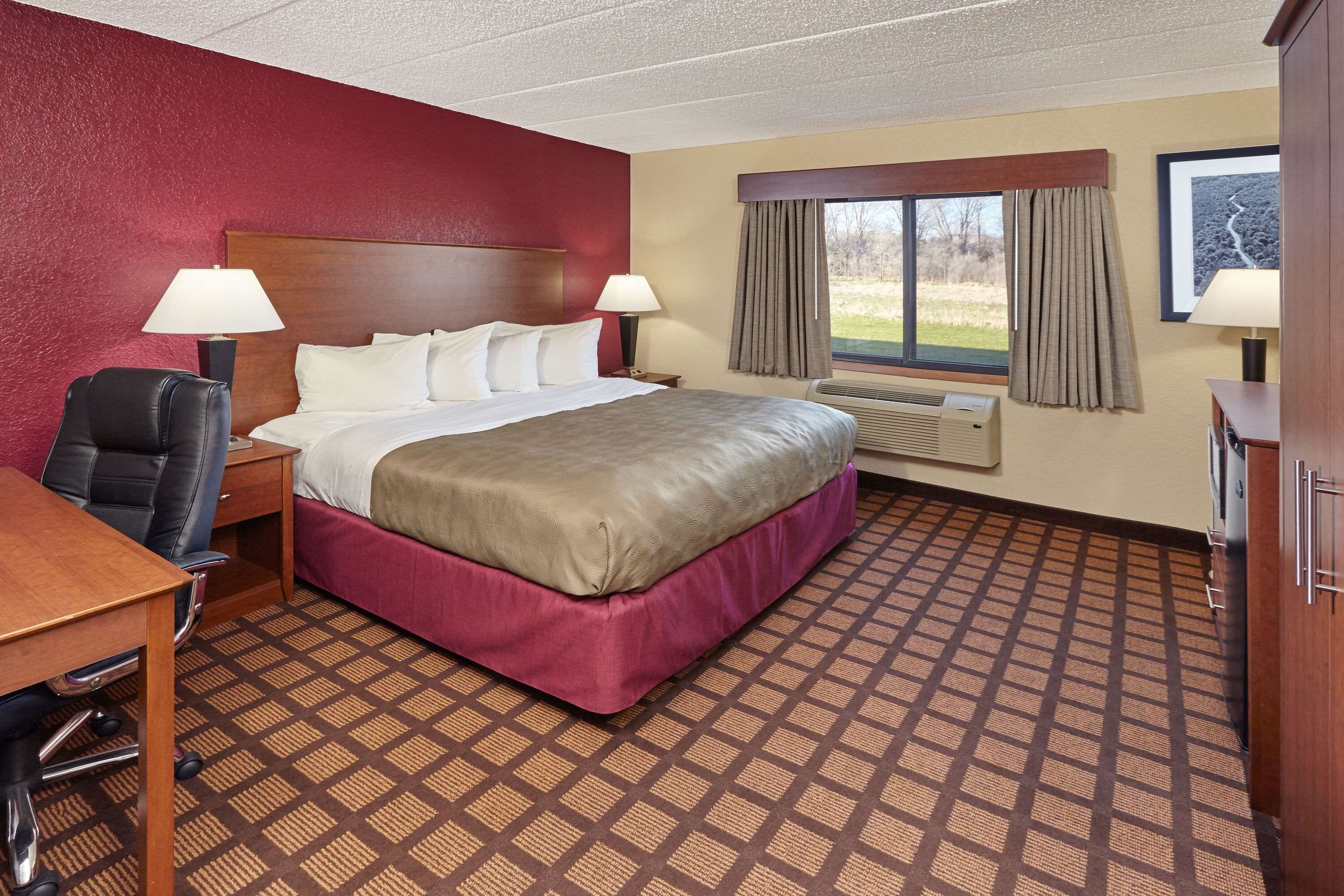 A hotel room with a large bed, two bedside tables with lamps, a desk with a chair, a wardrobe, and a window with curtains.