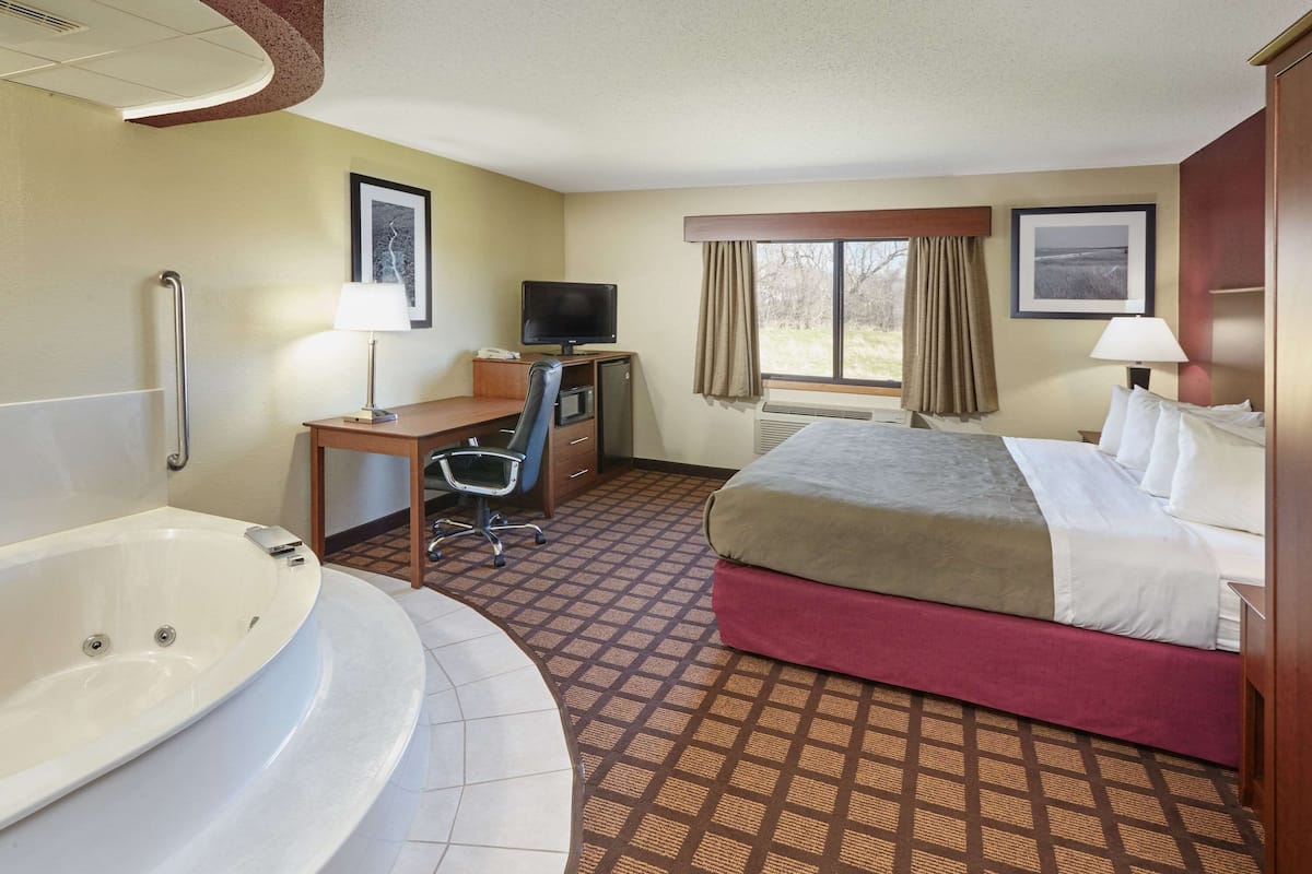 A hotel room with a large bed, a desk with a computer, a television, a bathroom with a bathtub, and a window with curtains.