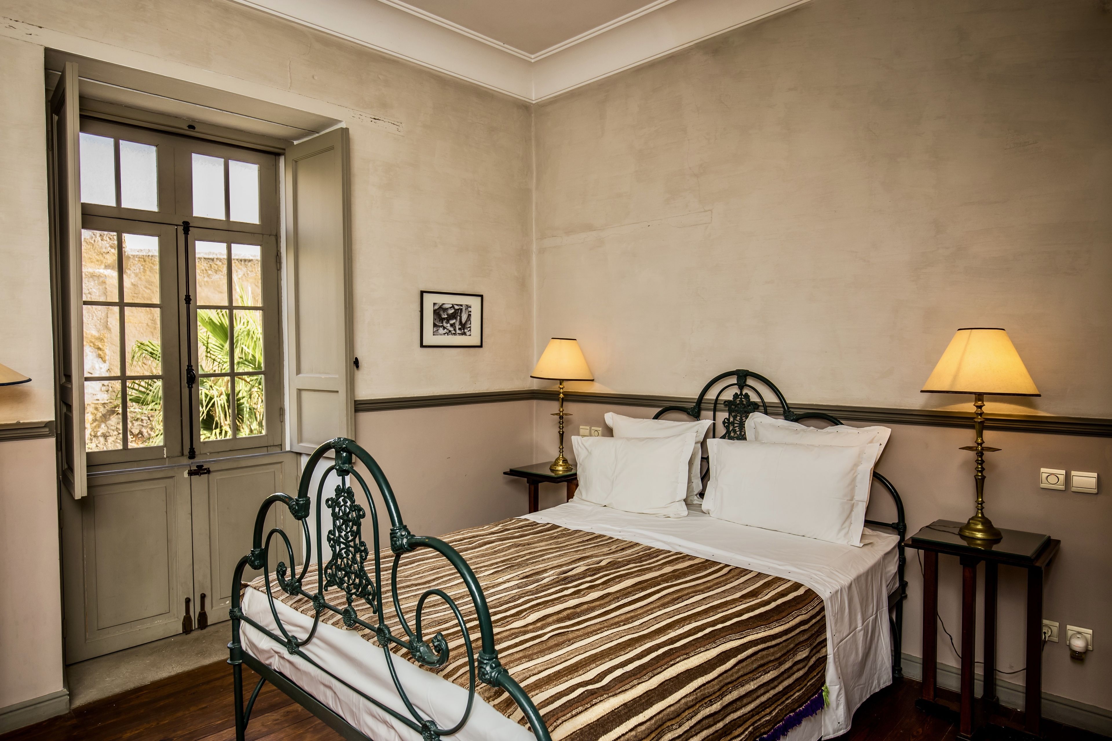 A bedroom with a bed, two bedside tables with lamps, a window with a view, and a framed picture on the wall.
